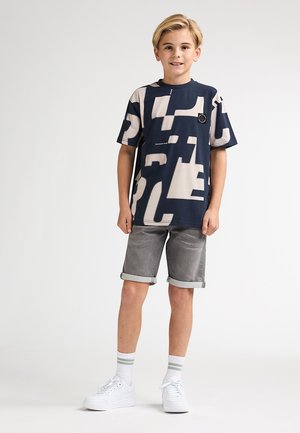 Tricou bleumarin cu modele abstracte bej, guler rotund. Asortat cu pantaloni scurți gri pliați și adidași albi, având șosete cu dungi.