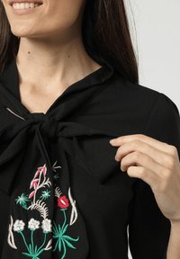 Top de tela negra con un cuello atado. Presenta bordados florales multicolores, destacando detalles en rojo, blanco y verde. Textura suave.