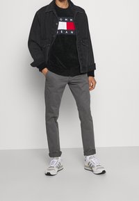 Veste en jean noire portée sur un pull noir avec un logo ; pantalon slim gris ; baskets grises et blanches avec des touches de bleu et de noir.