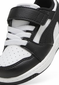 Svart-hvitt sneakers med teksturert skinnoverdel, perforert tå, hvite lisser og et borrelåsbelte for ekstra støtte.