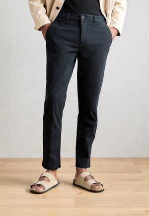 Homme portant un pantalon noir slim, des sandales beiges à bout ouvert avec boucles, une veste beige et une chemise noire, debout sur un sol en bois.