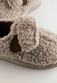 Beige fuzzy hjemmesko med tekstureret uldoverflade, mørkebrun stofdetalje og justerbar rem. Blødt materiale med en rund form.