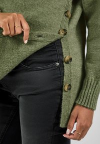 Cardigan verde a maglia con polsini a coste, caratterizzato da grandi bottoni marroni sul lato. Indossato sopra dei jeans neri, mostrando un tessuto con una miscela di texture.