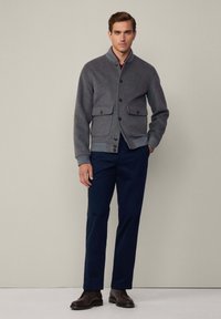 Chaqueta de lana gris con cierre de botones, dos bolsillos frontales y dobladillo acanalado. Combinada con pantalones azul marino y zapatos oscuros. Diseño sencillo.