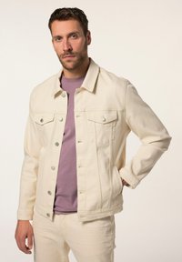 Chaqueta de mezclilla crema con botones plateados, dos bolsillos en el pecho y bolsillos laterales, llevada sobre una camiseta de color púrpura claro y pantalones a juego.