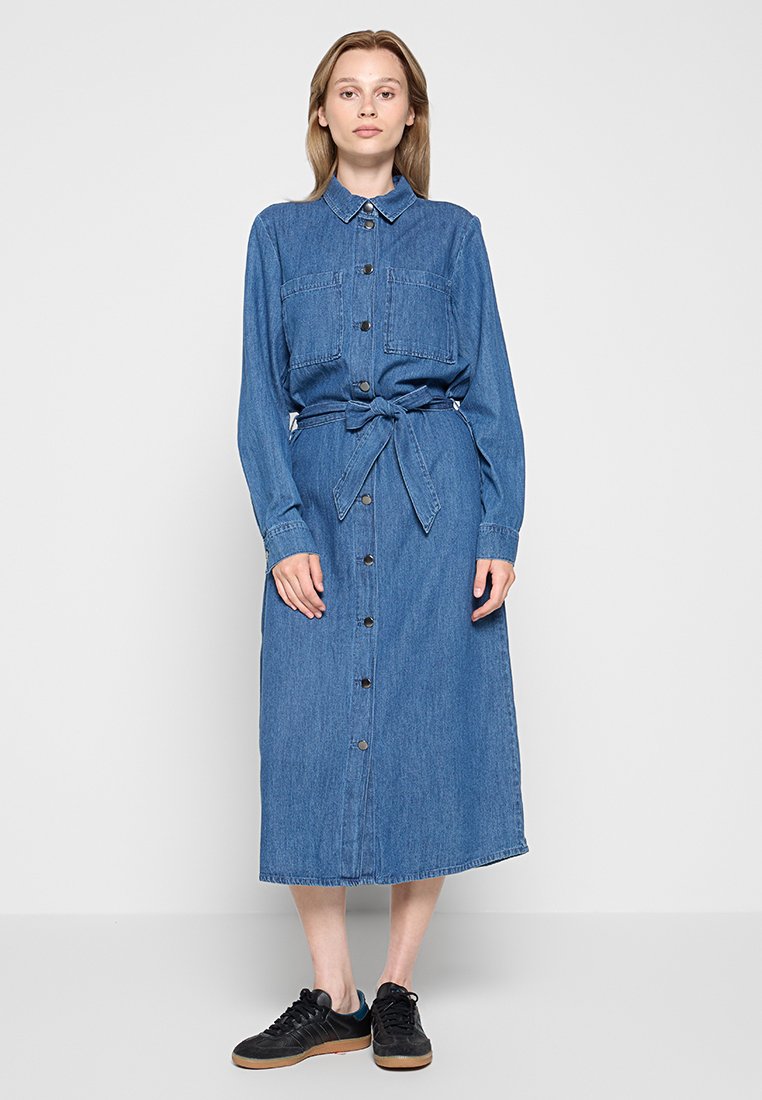 Robe chemise en denim, à manches longues, avec col, deux poches poitrine, boutonnière, ceinture à la taille, longueur mi-mollet, couleur bleue, portée avec des baskets noires.