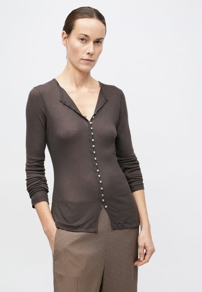 Barna hajú nő alacsonyan kontyba fogott hajjal, sötétbarna, szűk, hosszú ujjú felsőt visel kis gombokkal, és laza, taupe színű nadrágot.