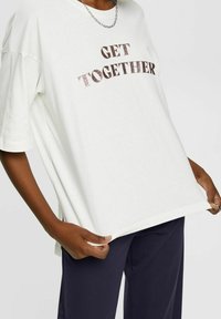 Camiseta blanca de tejido suave con el texto brillante "UNÁMONOS" en oro rosa. Presenta mangas cortas y un ajuste relajado. Combinada con pantalones azul marino.