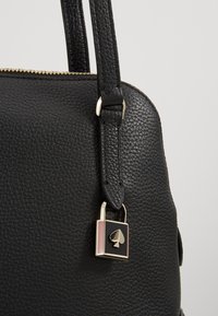 Sort læderhåndtaske med struktureret finish, guldfarvet hardware og en dekorativ låsevedhæng med et spar-ikon i pink og sort.