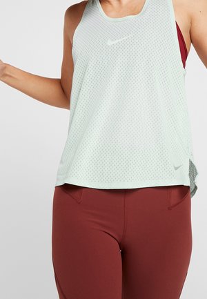 Frau trägt ein weißes ärmelloses Mesh-Sporttop über einem roten Sport-BH und roten, hoch taillierten Leggings vor neutralem Hintergrund.