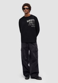 Svart sweatshirt med vit tryckt text, parat med svarta cargobyxor. Modell som bär mörka solglasögon och svarta skor. Minimalistisk design.