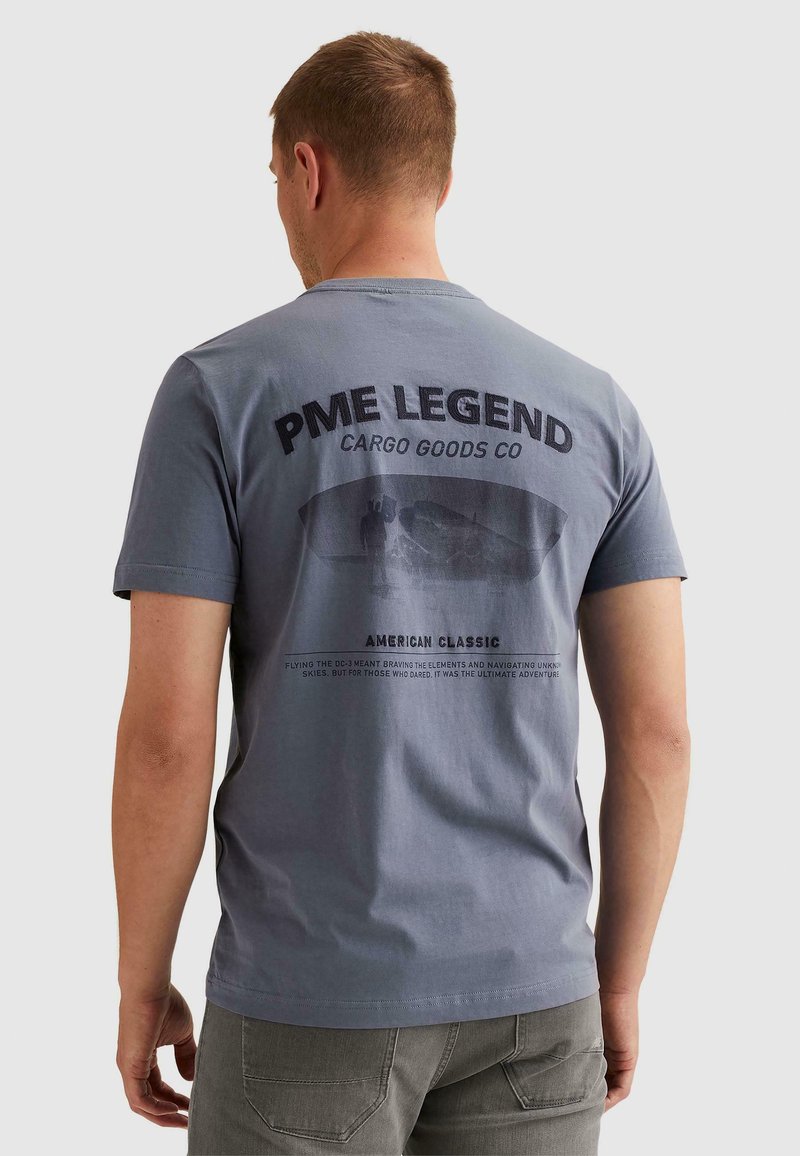 Άνδρας που φοράει γκρι T-shirt PME Legend με γραφικό αεροπλάνου φορτίου και κείμενο στην πλάτη, στέκεται μπροστά σε απλό φόντο.