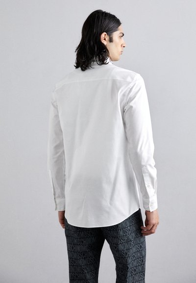 White long-sleeve shirt with a straight cut, featuring buttoned cuffs and a smooth texture, paired with patterned dark trousers.