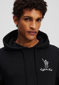 Schwarzer Hoodie aus Baumwolle mit einer Kängurutasche und einem weißen gestickten Design eines Kaffeeautomaten sowie dem Text "Café de Kol" auf der Brust.