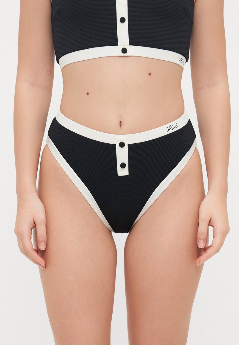 Svart teksturert bikini sett med en kort topp og høyt midje-bunn, begge med hvite detaljer og knapper, som viser en tett passform.