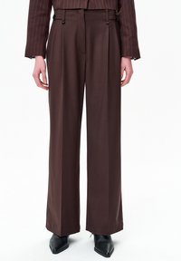 Persoon draagt bruine, hoge taille, wijde pijpen broek met voorplooien, gecombineerd met een bijpassend gestreept colbert en zwarte puntige schoenen.