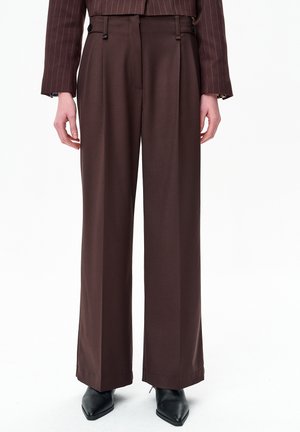 Persoon draagt bruine, hoge taille, wijde pijpen broek met voorplooien, gecombineerd met een bijpassend gestreept colbert en zwarte puntige schoenen.