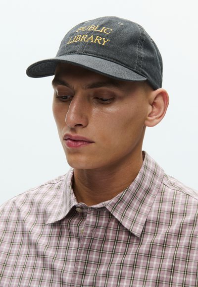 Γκρι καπέλο με τη φράση "PUBLIC LIBRARY" σε κίτρινα γράμματα. Φορεμένο με ανοιχτό ροζ καρό πουκάμισο, με πράσινες και λευκές γραμμές και γιακά με κουμπιά.
