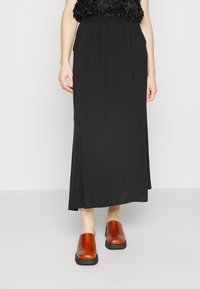 Zwarte, lange, vloeiende rok gemaakt van lichtgewicht stof, gecombineerd met bruine leren klompen met een dikke zwarte zool.