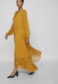 Vestido maxi amarelo mostarda, até os tornozelos, em tecido leve e fluido, com mangas longas e decote redondo. Sapatos de salto pretos.