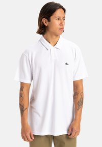 Weißes Polohemd aus Baumwolle mit einem schwarzen gestickten Logo auf der Brust, klassischem Kragen und kurzen Ärmeln.