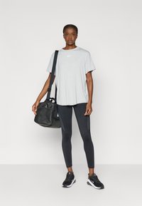 Γκρι μπλούζα Nike, μαύρο κολάν με υφασμάτινα πάνελ, κρατώντας μια μαύρη τσάντα duffel, φορώντας μαύρα και λευκά αθλητικά παπούτσια. Απλός σχεδιασμός, αθλητική εφαρμογή.