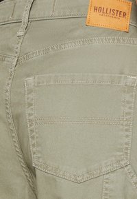 Khaki džíny se slim fit střihem, s viditelnou zadní kapsou, kontrastním šitím a hnědou koženou značkovou nášivkou na pase.