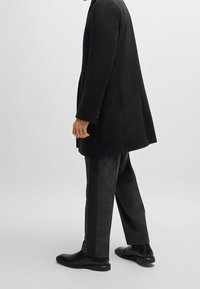 Cappotto lungo nero con una vestibilità sartoriale, indossato sopra pantaloni neri con un motivo a diamante, e scarpe in pelle nere.