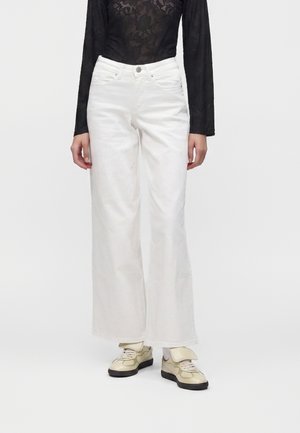 La personne porte un pantalon blanc à jambes larges, des baskets beiges avec des semelles noires, et un haut en dentelle noire à manches longues avec des motifs floraux.