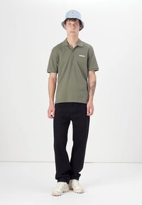 Olivengrønn polo-skjorte med krave, svarte bukser, lys blå bøttehatt og beige joggesko. Inneholder diskret merkevare på skjorten.