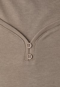 Tessuto di cotone color beige con design a scollo a V, caratterizzato da due bottoni marroni sulla scollatura e una texture liscia. Dettaglio di cuciture visibili.