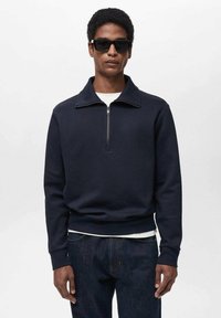 Tmavomodrá mikina s polovičným zipsom a golierom, vyrobená z mäkkej látky. Má pružné manžety a spodný lem, nosená cez bielu košeľu, v kombinácii s tmavými džínami.