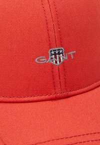 Căciulă de baseball roșie, dintr-un material texturat, cu un logo brodat având un design în formă de scut. Accente cusute pe margine și pe coroana căciulii.