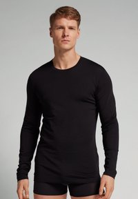 Schwarzes Langarmshirt aus weichem Stoff, mit Rundhalsausschnitt und taillierter Passform. Getragen über schwarzen Shorts, glatte Textur ohne Muster.