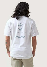 Camiseta de algodón blanca con un diseño gráfico en la parte posterior que presenta un símbolo de ancla y una línea ondulada en azul claro. Mangas cortas y un ajuste relajado.