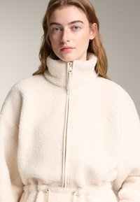 Jakna iz kremastega fliša z visokim ovratnikom, zadrgo po celotni sprednji strani in vrvico za prilagajanje pasu. Ima mehko, teksturirano in udobno teksturo.