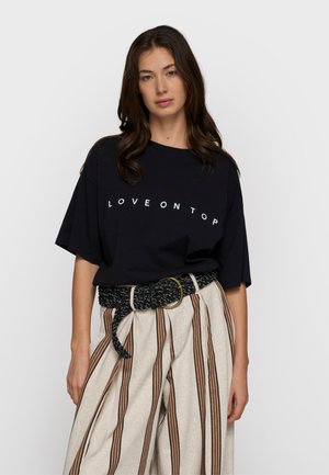 Kobieta o długich ciemnych włosach, ubrana w czarną koszulkę z napisem "LOVE ON TOP" oraz beżowe spodnie z pionowymi brązowymi paskami i czarnym paskiem.