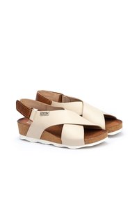 Beżowe skórzane sandały z krzyżującymi się paskami, brązowy akcent na pięcie, korek w śródstopiu i biała podeszwa. Zamknięta tylnia część dla pewnego dopasowania.