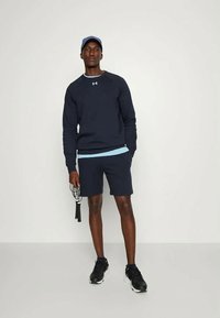 Marineblå hettegenser med lyseblå detaljer, kombinert med marineblå shorts. Modellen holder et hoppetau og har på seg svarte joggesko og en caps.