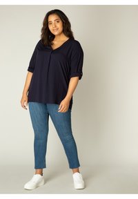 Marineblauw T-shirt met korte mouwen en een V-hals, voorzien van plooidetails, gecombineerd met lichtblauwe slim-fit jeans en witte sneakers, wat zorgt voor een casual outfit.