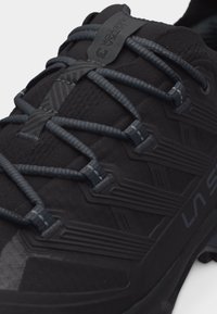 Must La Sportiva tossuga lähedalt, millel on halli mustriga paelad ja tekstuurse sünteetilise pealismaterjaliga pealiskate.