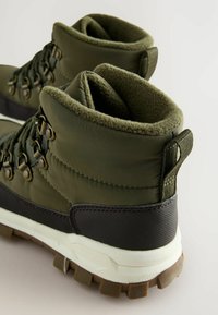 Grüne wasserdichte Wanderschuhe mit einem gepolsterten Schaft, Metallösen für die Schnürsenkel und einer strukturierten schwarzen Gummisohle mit braunem Profil.