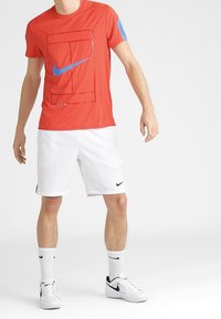 Röd tränings T-shirt med blå Nike-logga, korta ärmar, vita shorts, svart Nike-accent på shortsen, vita strumpor och svarta sportskor.