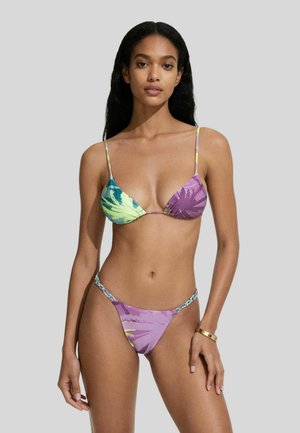 Donna con capelli lunghi e scuri che indossa un bikini viola e verde con fantasia e spalline sottili, posando davanti a uno sfondo grigio uniforme.