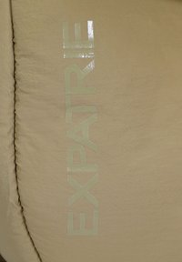 Surface en tissu beige avec le mot "EXPATRIE" imprimé verticalement en lettres brillantes et réfléchissantes.