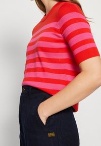 Punainen ja vaaleanpunainen raidoitettu neulepaita, jossa on lyhyet hihat, pyöreä pääntie ja rento istuvuus. Yhdistetty tummansinisien samettihousujen kanssa.