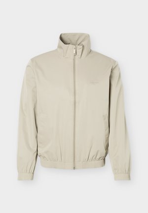 Veste légère beige avec col montant, fermeture éclair à l'avant, poignets et ourlet élastiques, dotée de poches latérales et d'un logo discret sur la poitrine.