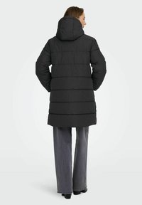 Melnā pūkainā jaka ar kapuci, ar quilted dizainu un horizontālām svītrām. Apvienota ar platām pelēkām biksēm un zābakiem.