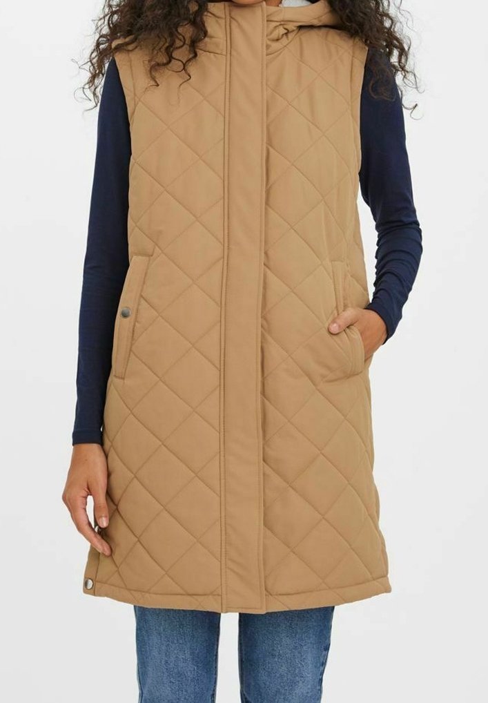 Femme portant une veste matelassée sans manches beige sur une chemise bleu marine à manches longues et un jean bleu, avec une main dans la poche de la veste.