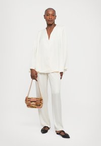 Blusa de seda blanca con escote en V y mangas largas, combinada con pantalones blancos de pierna recta. Sosteniendo un bolso de mano tejido multicolor. Zapatos planos negros.